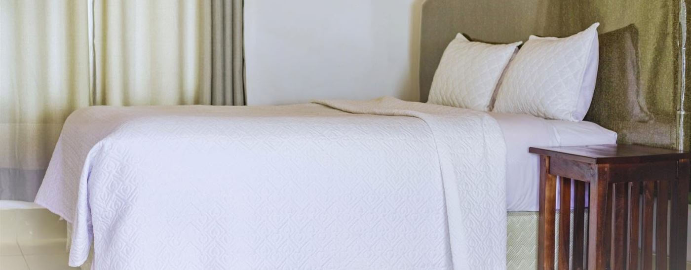 A Harare room with a made white bed, wooden side table and a green padded headboard
