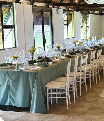 Long tables dressed in sage green cloth with white chairs and yellow flowers under a thatched roof
